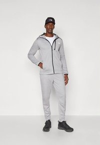 Άνδρας που φοράει γκρι φούτερ με φερμουάρ και αντίστοιχο παντελόνι φόρμας, μαύρο καπέλο και μαύρα παπούτσια πεζοπορίας, στέκεται με το ένα χέρι στην τσέπη.