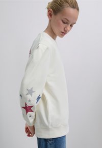 Valkoinen collegepaita, jossa on pitkät hihat ja vasemmassa hihassa punainen tähti, hopeinen tähti ja sininen salamanisku. Resorit hihansuissa, rento istuvuus.