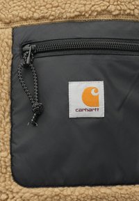 Chaqueta de forro polar con un bolsillo de nailon negro con cierre de cremallera, forro polar texturizado en color beige y una etiqueta blanca con un logo naranja.