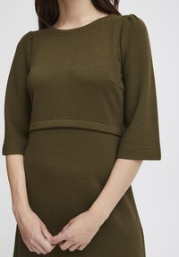 Olivenfärgad klänning med en texturerad, strukturerad tyg, med rund halsringning och trekvartsärmar samt en figurnära midja.