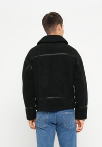 Schwarz strukturierte Fleecejacke mit breitem Kragen, versehen mit Lederakzenten entlang der Nähte. Der Rücken hat verstellbare Riemen und eine glatte Oberfläche.