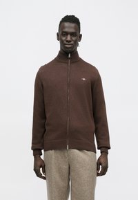 Brūns rāvējslēdzēja džemperis ar augstu apkakli, adīts tekstūra un logotipa detaļa kreisajā krūtīs; kopā ar gaismas krāsas teksturētām biksēm.