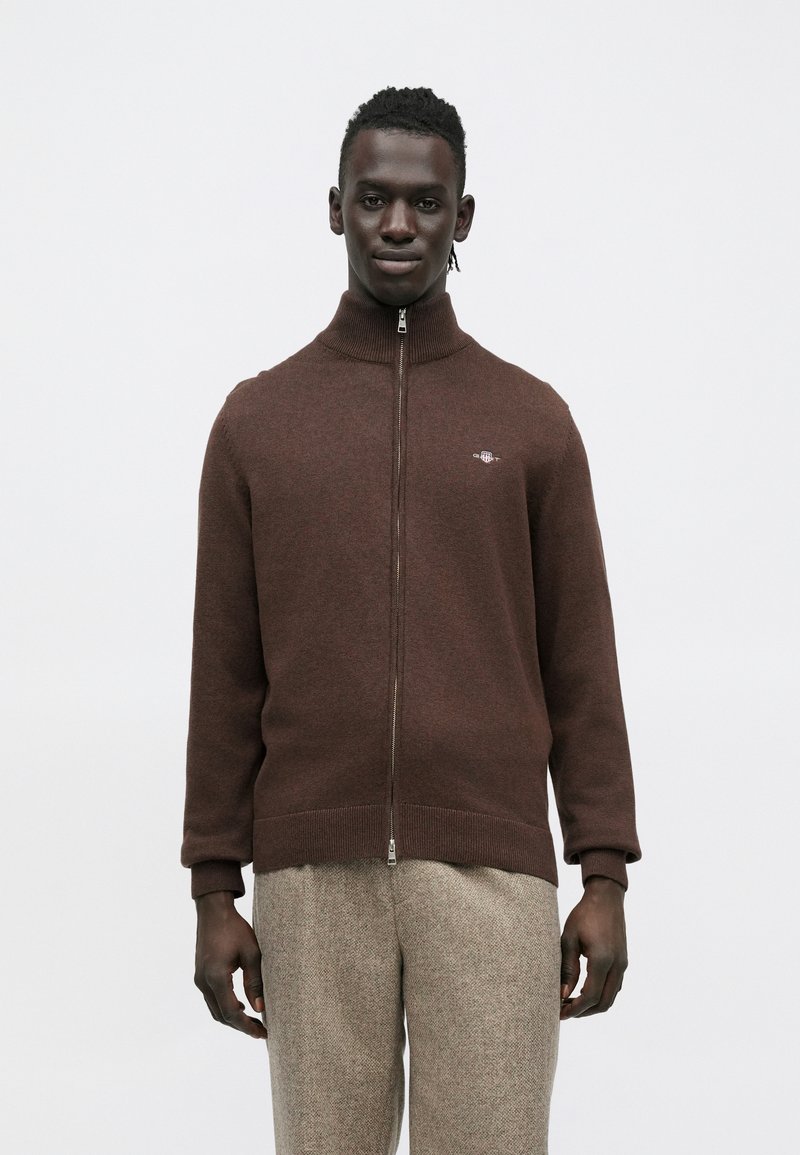 Brūns rāvējslēdzēja džemperis ar augstu apkakli, adīts tekstūra un logotipa detaļa kreisajā krūtīs; kopā ar gaismas krāsas teksturētām biksēm.