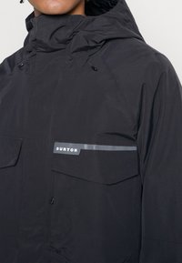 Schwarze wasserdichte Jacke mit hohem Kragen, ausgestattet mit zwei großen Vordertaschen und einem reflektierenden Streifen. Das Material wirkt glatt und strapazierfähig.
