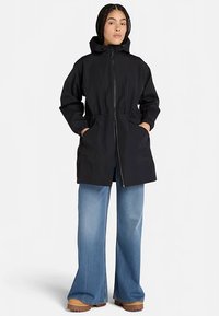 Svart vattentät jacka med huva, elastisk dragsko i midjan, dragkedja fram och två sidofickor; kombinerad med vida jeans.