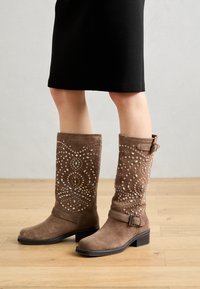 Botas hasta la rodilla de ante marrón con patrones decorativos de tachuelas y una correa ancha con hebilla metálica. Tacón bajo apilado, textura suave.