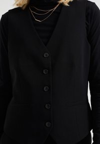 Svart väst med enåtsittande design, som har sex framknappar, två sidofickor och en slät, vävd yta.