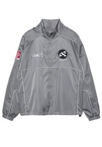 Sivá športová bunda s celým zipsom, text USA 91 na ľavej strane hrudníka, športové logo na pravej strane hrudníka a červená nášivka na ľavom rukáve.
