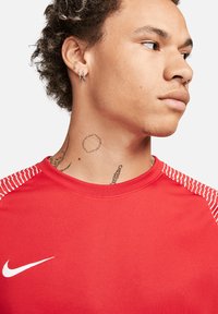 Rotes Sportshirt mit weißem Nike-Swoosh-Logo, mit gestreiften Akzenten an den Schultern. Der Stoff wirkt glatt und enganliegend.