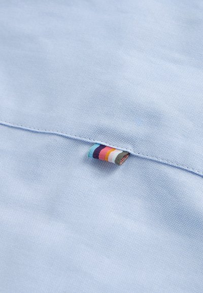 Šviesiai mėlyna medžiaga su tekstūruotu audiniu, kuriame įsiūtas mažas ženkliukas su daugiaspalvėmis juostelėmis.