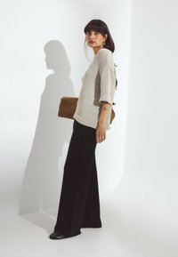 Femme aux longs cheveux foncés portant un pull beige, un pantalon noir à jambes larges, tenant une pochette marron, debout devant un fond blanc projetant une ombre.