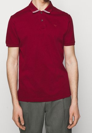 Burgundské polo tričko s dvouknoflíkovou légou, límcem s bílými a červenými pruhy a jemným logem na hrudi. Tkanina se zdá být hladká a prodyšná.