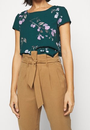 Blusa floreale turchese con maniche corte e scollo a girocollo, abbinata a pantaloni beige a vita alta con cintura annodata e fronte plissettato.