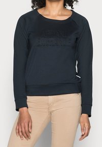 Černá mikina s kulatým výstřihem, raglánovými rukávy a vyraženým logem "ARMANI EXCHANGE" na přední straně. Měkká tkanina a vypasovaný pas.