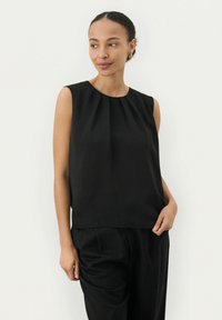 Top nero senza maniche con scollatura plissettata, realizzato in tessuto liscio. Abbinato a pantaloni neri a gamba larga, con un fit rilassato.