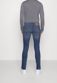 Sinisiä farkkuja, joissa on kapea malli, takataskut ja ruskea nahkalaatta vyötäröllä, yhdistettynä harmaaseen pitkähiharaiseen paitaaan.