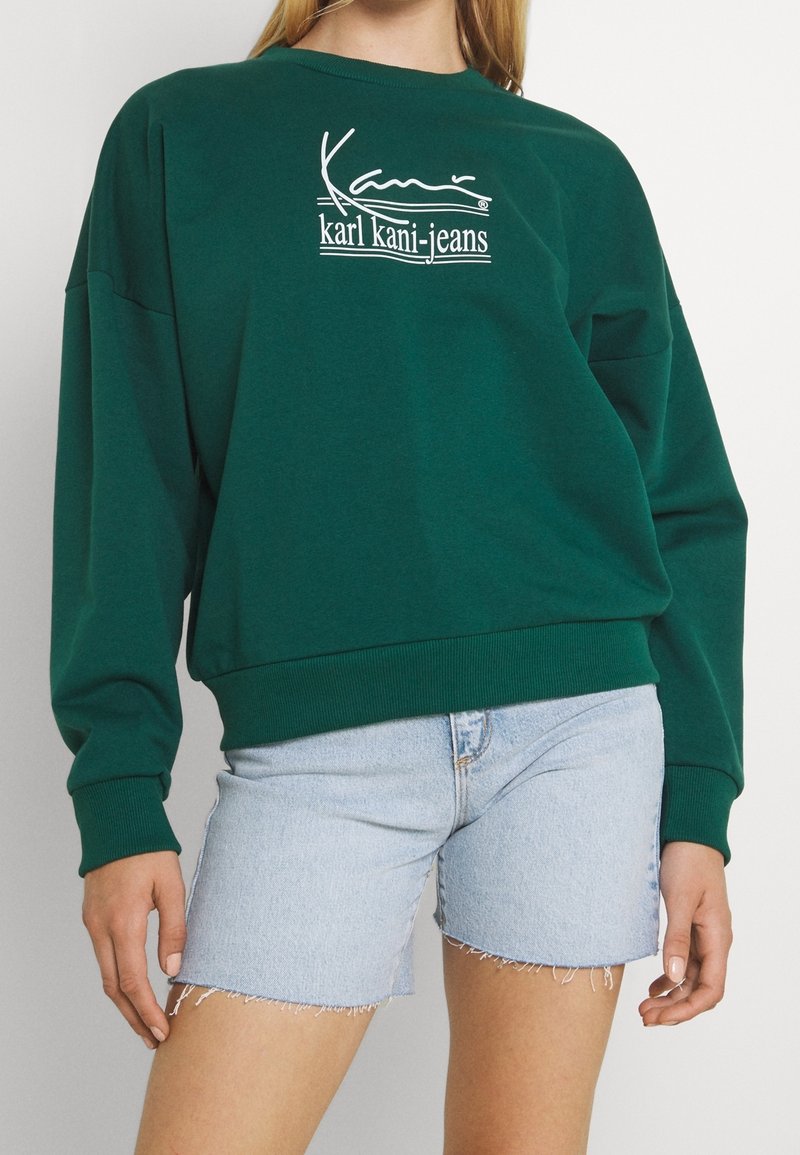 Sweatshirt en coton vert avec un col rond, arborant un logo blanc imprimé sur la poitrine. Associé à un short en denim bleu clair coupé.