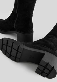 Zwarte suede kniehoge laarzen met een blokhak en een getextureerde rubberen zool met een zigzagpatroon. Zijrits voor gemakkelijke aan- en uittrekken.