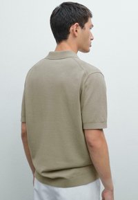 Polo tričko s krátkým rukávem v světle taupe, vyrobené z hladkého materiálu. Má klasický límec a žebrovaný lem, s čistým, minimalistickým designem.