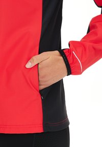 Giacca sportiva rossa e nera con tasca laterale con zip, polsini elasticizzati e un motivo di tessuto testurizzato. Accenti riflettenti visibili.