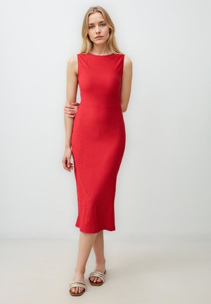 Red ribbed midi dress with a fitted silhouette, round neckline, and side slits. Model wears neutral slide sandals on a textured surface.