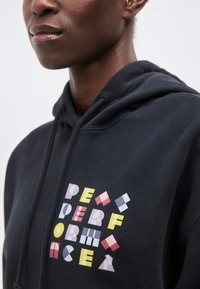 Κοντινό πλάνο ενός ατόμου που φοράει μαύρη κουκούλα με πολύχρωμα γεωμετρικά γράμματα που σχηματίζουν αφηρημένο σχέδιο στο στήθος.