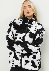 Veste en polaire avec un imprimé vache noir et blanc, col montant, fermeture éclair complète et poches latérales. Texture douce et coupe décontractée.