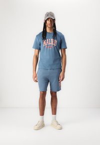 Blauwe katoenen T-shirt met 'Malibu'-print, bijpassende blauwe shorts en beige sneakers. Accessoires omvatten een grijze pet met kleurrijke ontwerpen.