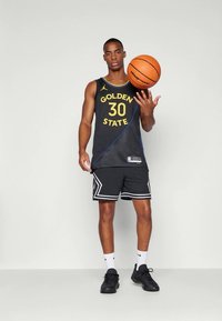 Vyras, vilkintis Golden State krepšinio marškinėlius, suktis krepšinį ant vieno piršto prieš paprastą foną.