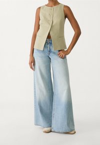 Helles grünes ärmelloses Oberteil mit Frontknöpfen, kombiniert mit weit geschnittenen, verblassten Jeans. Sanfte Texturen und strukturiertes Design.