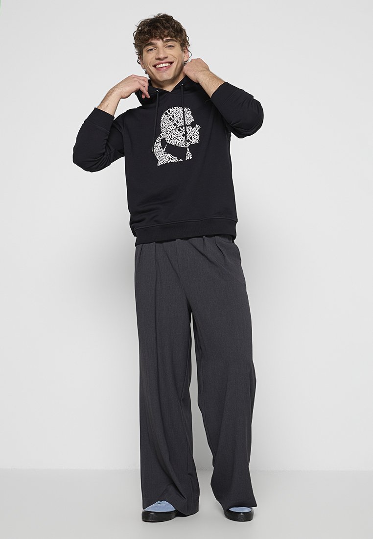Sweat à capuche noir avec un design graphique blanc, associé à un pantalon gris ample. La tenue présente une coupe décontractée et une texture de tissu douce.