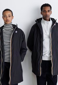 Sorte overtøjjakker med kontrastfarvede hvide lynlåse på begge modeller. Den ene har en stribet turtleneck på; den anden har en grå zip-up top.