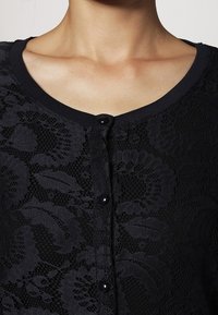 Cardigan in pizzo nero con scollo rotondo, chiusura a bottone e intricate decorazioni floreali su tutto il tessuto, rifinito con una delicata bordatura a coste.