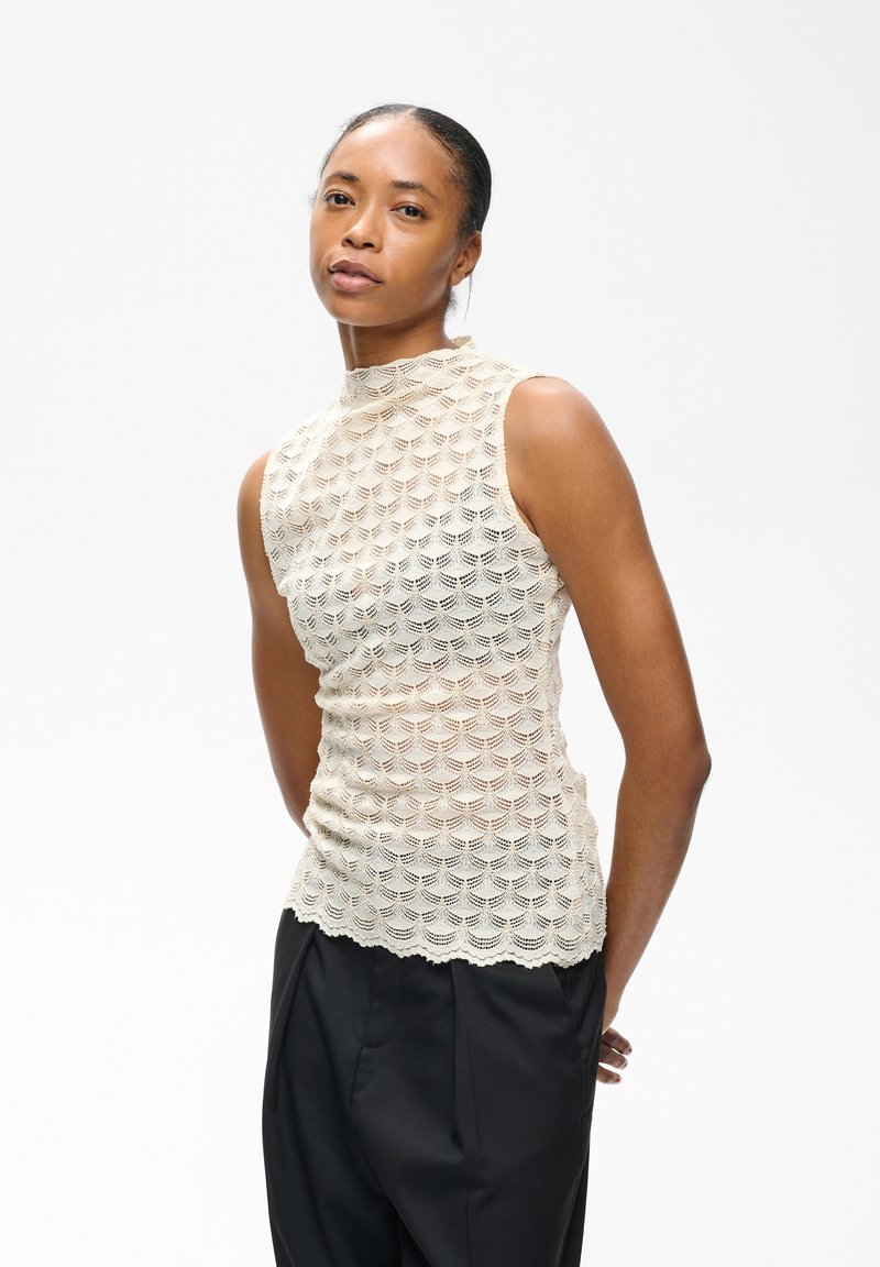 Femme aux cheveux attachés portant un haut en dentelle crème sans manches et un pantalon noir, posant avec les mains derrière le dos sur fond blanc.