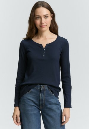 Femme portant une chemise henley à manches longues bleu marine légèrement rentrée dans un jean bleu délavé, se tenant contre un fond clair.