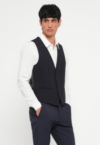 Vest negru cu cinci butoni și două buzunare frontale, purtat peste o cămașă albă. Țesătură netedă cu un design slim fit și fără modele vizibile.