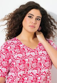 Halenka s výstřihem do V v jasně růžové barvě s bílým botanickým vzorem; krátké rukávy; měkká, strukturovaná tkanina; modelka předvádějící náušnice a účes.