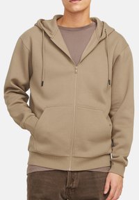 Hættejakke med lynlås i taupe, med en kengurulomme, snore og ribbet manchetter. Laget over en brun T-shirt.