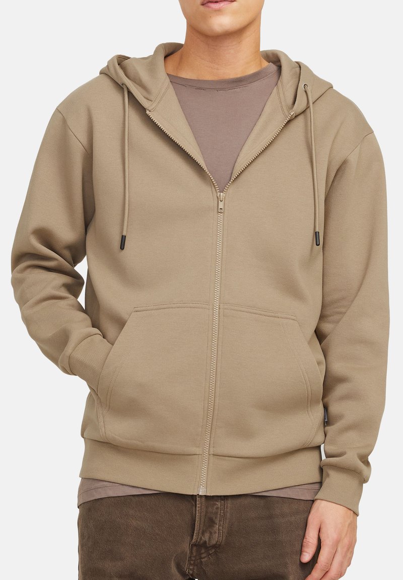 Hættejakke med lynlås i taupe, med en kengurulomme, snore og ribbet manchetter. Laget over en brun T-shirt.