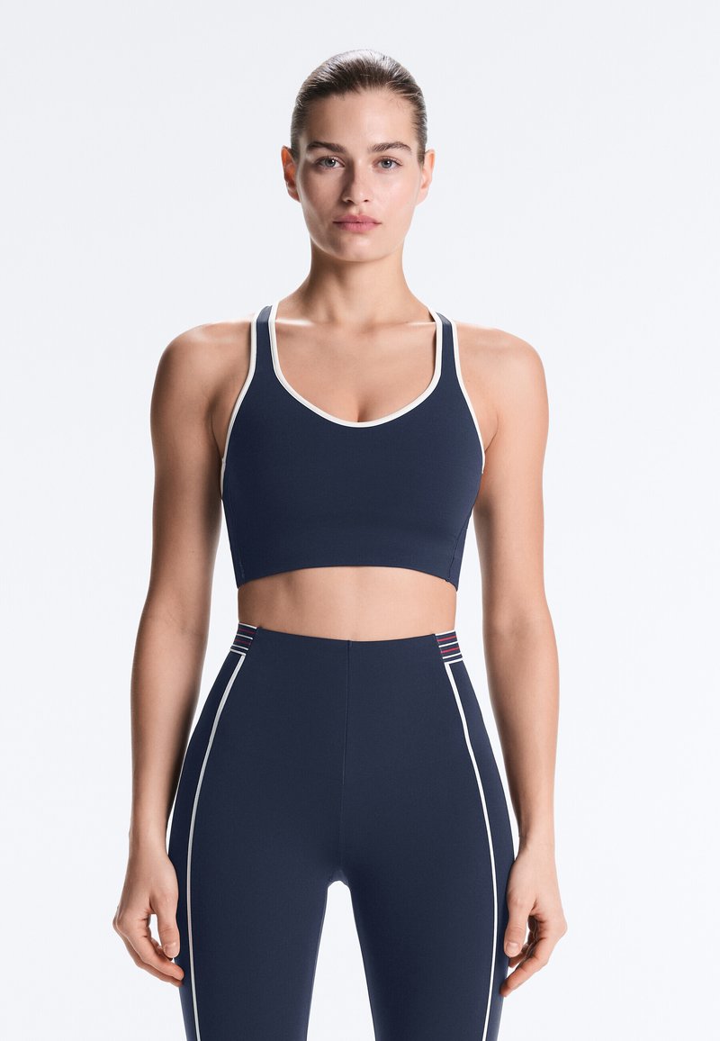 Femme debout, faisant face à l'avant, portant un soutien-gorge de sport bleu marine et un legging taille haute avec des bordures blanches et des accents de bandes rouges.