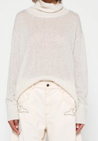 Maglione a punto di maglia beige chiaro con collo alto, caratterizzato da una vestibilità comoda e un tessuto testurizzato, abbinato a pantaloni color crema con cuciture decorative.