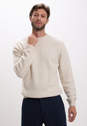 Maglione lavorato a maglia color crema con motivo a treccia, dotato di colletto tondo e polsini a costine. Indossato sopra pantaloni scuri, mostrato su uno sfondo bianco.