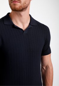 Tumši zilā krāsā īsām piedurknēm polo krekls ar ribotu tekstūru un klasisku apkakli, kas aprīkota ar trīs pogu aizdari.