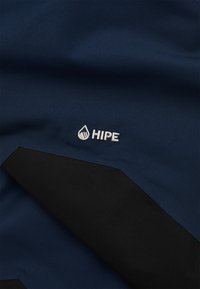 Marineblåt stof med sort struktureret overlay; har en hvid broderet "HIPE" logo. Glat tekstur med et strømlinet design.
