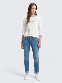 Chemise blanche à manches longues avec "Tom Tailor" imprimé en noir ; associée à un jean bleu et des baskets blanches ; coupe décontractée, avec les manches du jean retroussées.