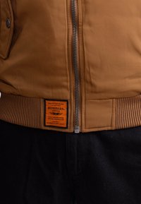 Bruine bomberjack met een geribbelde gebreide zoom, ritssluiting en zwarte drukknopzakken. Heeft een oranje label aan de onderkant van de voorkant.