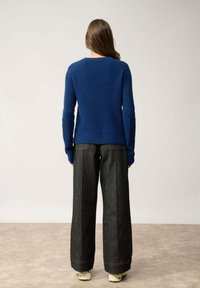 Pull en tricot côtelé bleu foncé à manches longues, associé à un jean en denim large et foncé. Le décor présente un arrière-plan neutre.