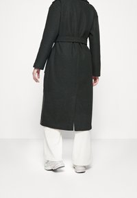 Abrigo largo verde oscuro con un cinturón, que presenta una abertura trasera y mangas largas, combinado con pantalones anchos de color claro y zapatillas.