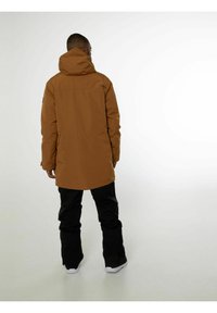 Parka imperméable marron avec capuche, coupe mi-longue, pantalon noir et baskets blanches. Marquage minimal à l'arrière.