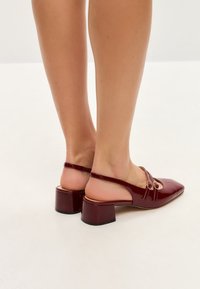 Burgundi színű lakk slingback magassarkú cipők, négyzet alakú orral, két keresztezett pánttal és vastag cipőtalppal. A felület fényes, aranyszínű fém díszítésekkel.
